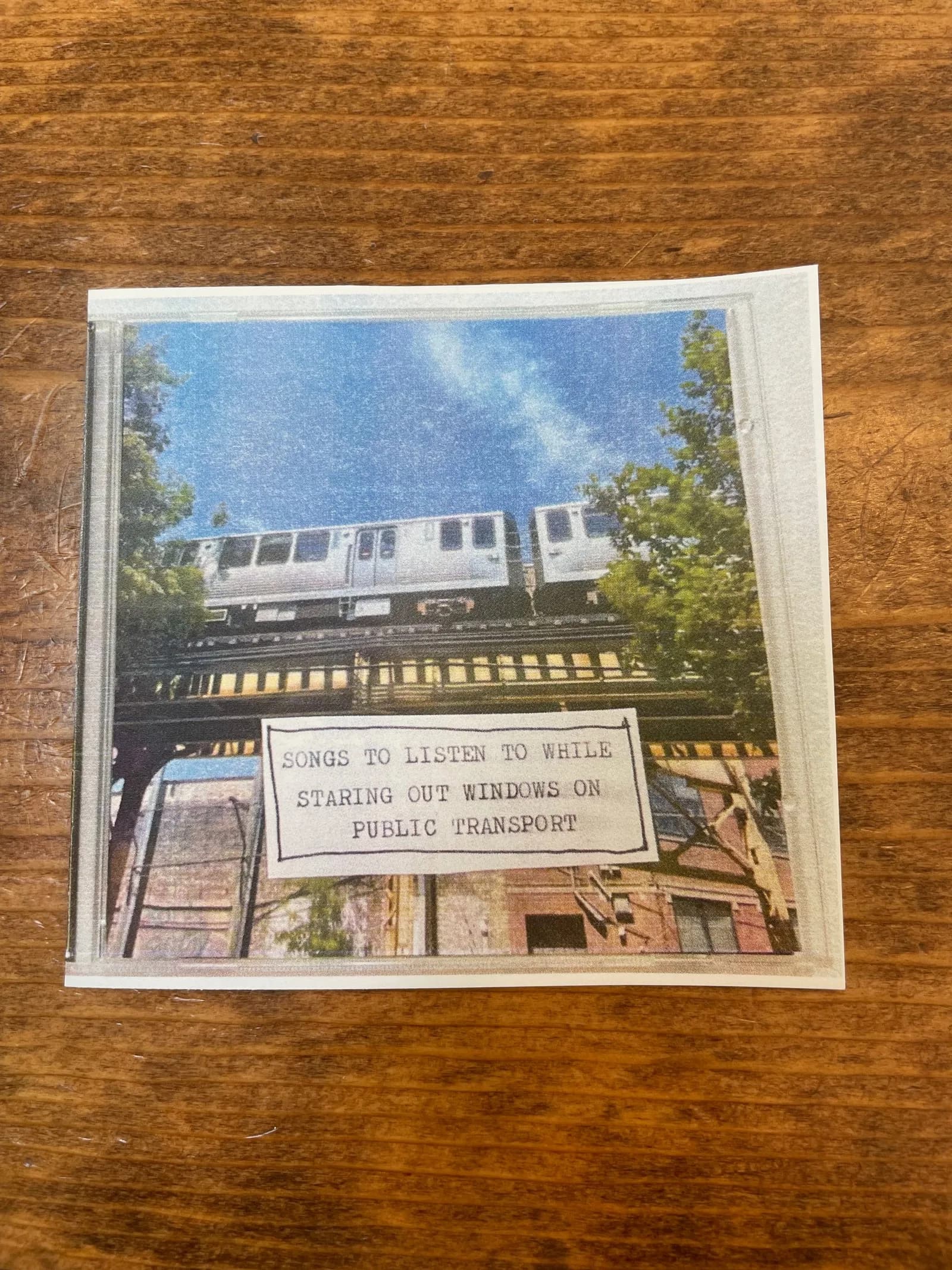 The zine in a CD case on a wooden table: an elevated train over the tracks, with a typed label reading SONGS TO LISTEN TO WHILE STARING OUT WINDOWS ON PUBLIC TRANSPORT.