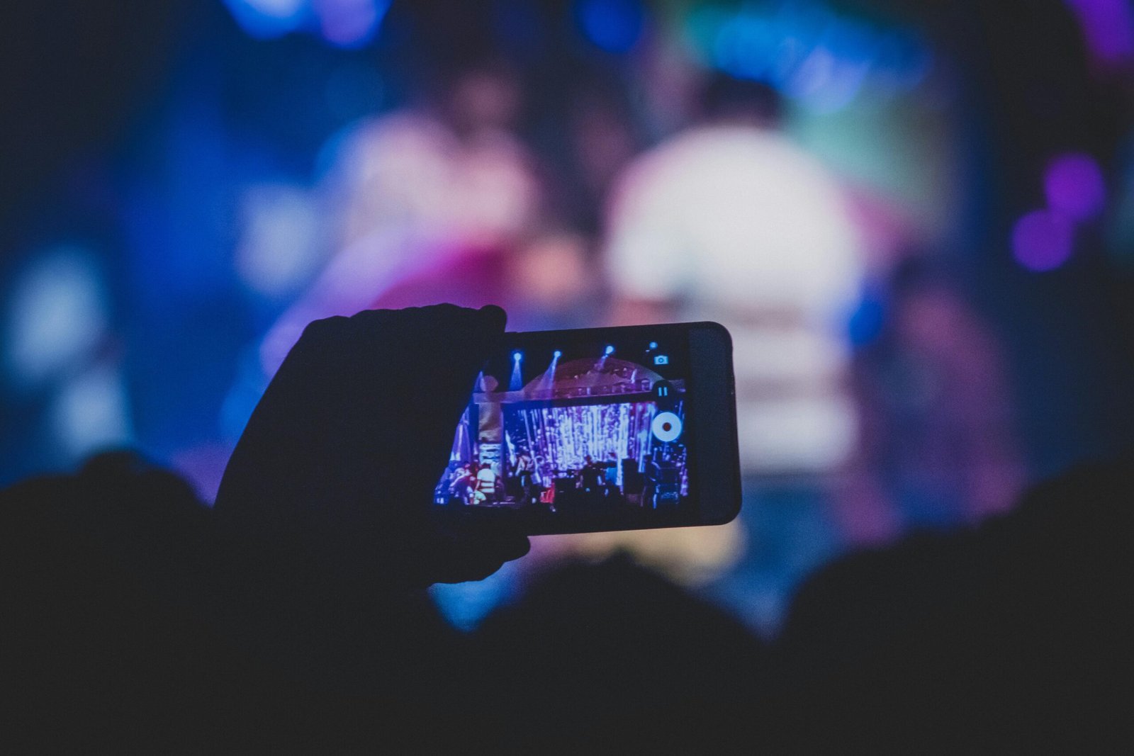 A hand holds a smartphone recording a live concert with vibrant lights.